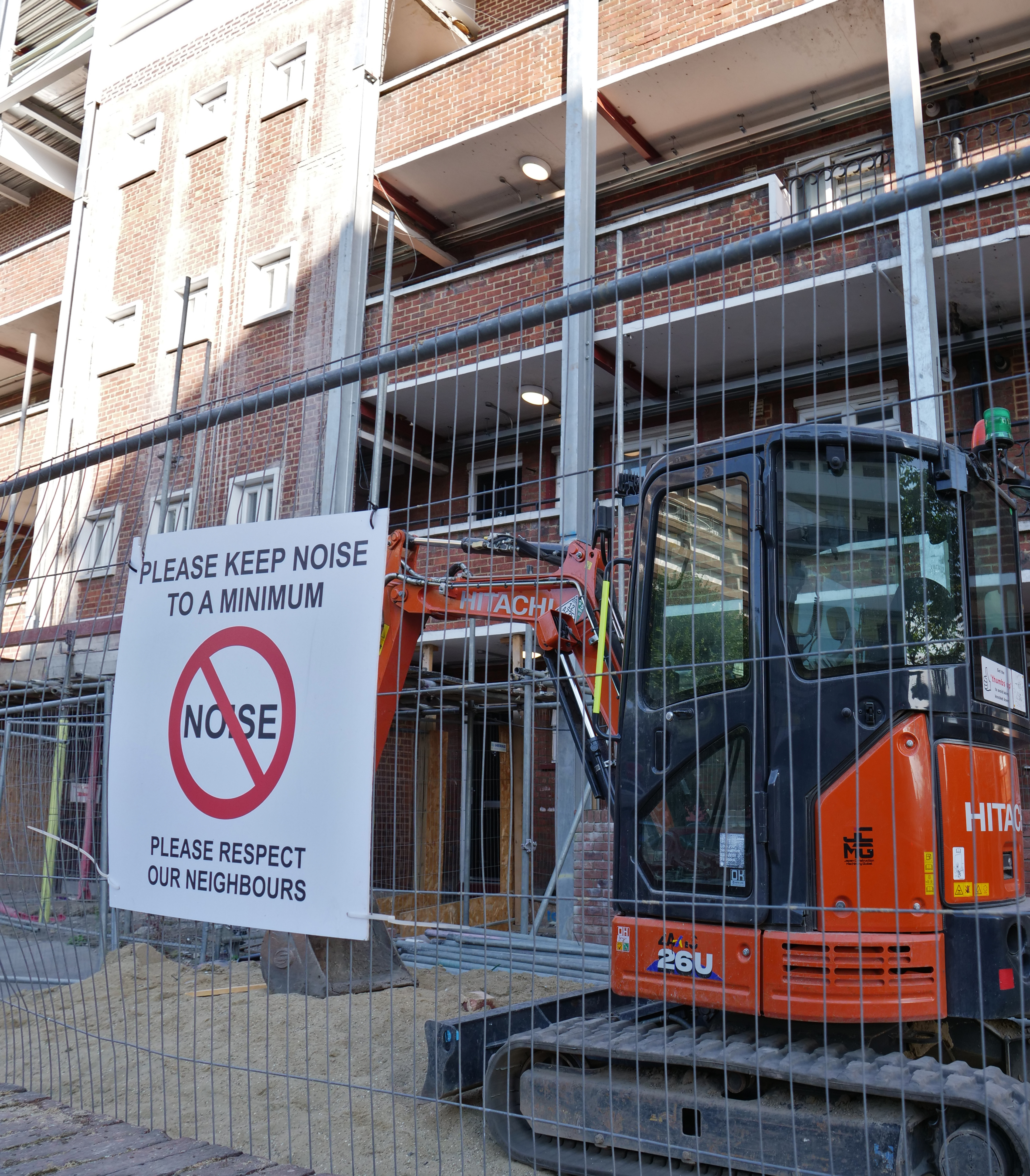 A sign attached to a Heras fence admonishing passers by to "PLEASE KEEP NOISE TO A MINIMUM". Behind the fence is a Hitachi mini digger on top of a pile of sand.