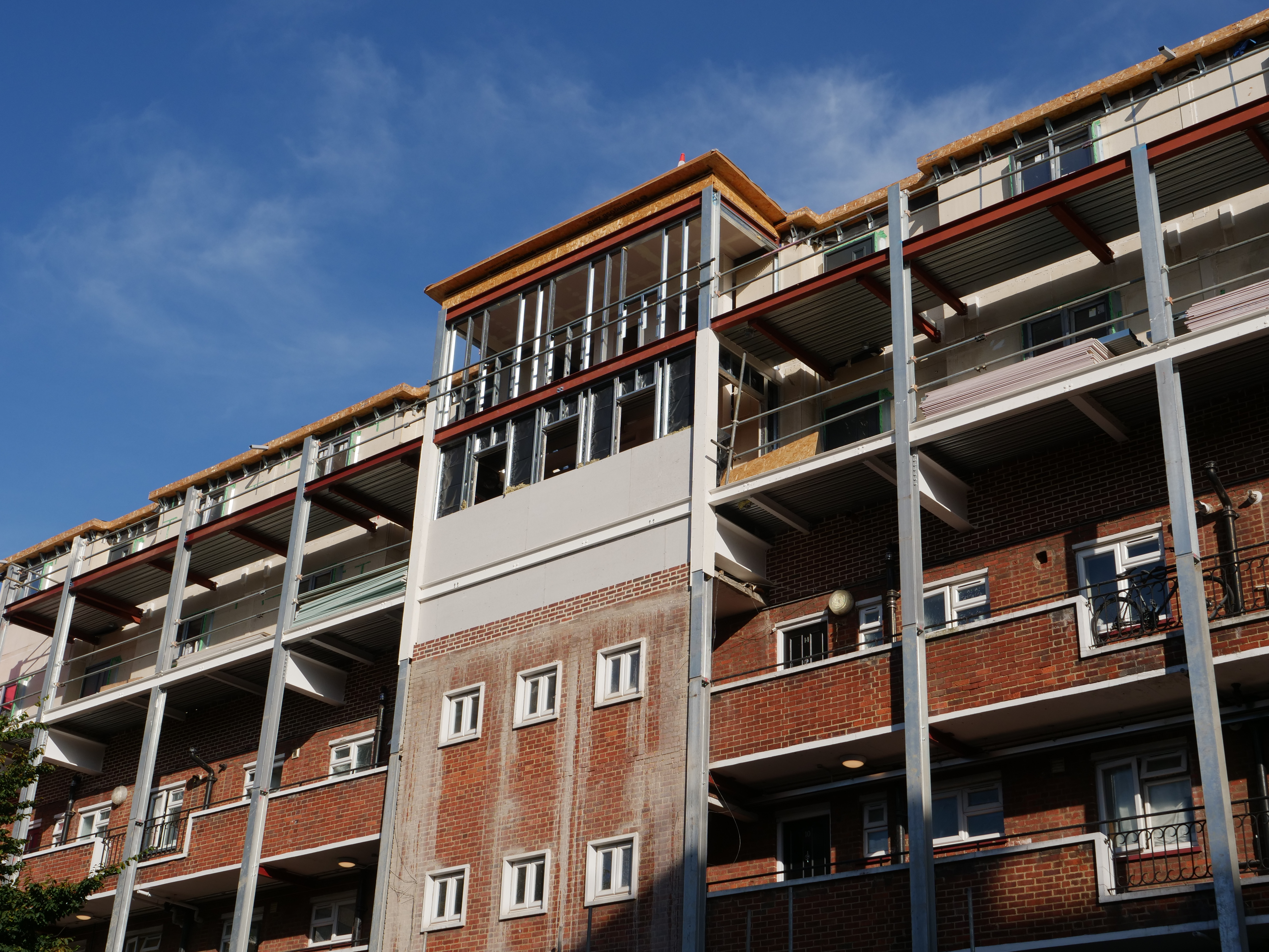 Antony House with the new block of flats on top partially complete. The bare walls of the prefabricated modules can be seen without any cladding. The new flats are supported by a series of strong steel beams, supported by silver-coloured galvanised steel columns. The facade of Antony House is stained with cement.